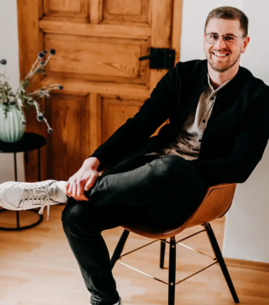 Lukas Kölzer, Steuerberater und Inhaber der Kölzer Steuerberatungsgesellschaft sitz locker auf einem Stuhl in modernem Outfit und lächelt freundlich. Im Hintergrund ist eine Pflanze und eine Türe der Büroräumlichkeiten der Steuerberatung Kölzer zu sehen.